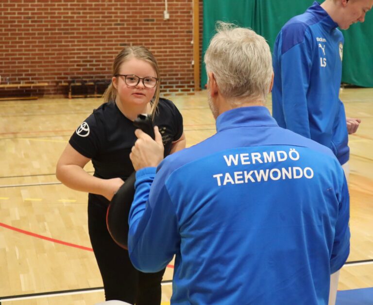 En flicka tränar taekwondo i en idrottshall tillsammans med en instruktör från Wermdö Taekwondo. Hon bär glasögon och en svart t-shirt med svenska paralympiska kommitténs logotyp, och ser fokuserad ut inför ett slag mot den mitts som instruktören håller upp. Instruktören har på sig en blå jacka med texten “Wermdö Taekwondo” på ryggen. En annan person i blått träningsställ syns i bakgrunden.